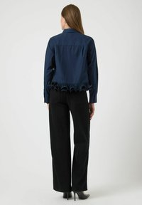 Vrouw die met haar rug naar ons toe staat, gekleed in een marineblauwe blouse met een gerimpelde zoom, zwarte wijde broek en zwarte hoge hakken op een witte achtergrond.