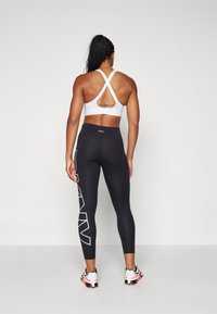 Black leggings with white logo print, paired with a white racerback sports bra, revealing a tattoo on the back, worn with athletic shoes.