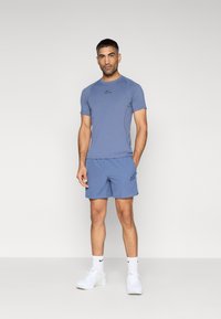 Αθλητική μπλούζα κοντομάνικη σε ανοιχτό γαλάζιο με μαύρο λογότυπο, συνδυασμένη με σορτς στο ίδιο χρώμα. Το σύνολο ολοκληρώνουν λευκά αθλητικά παπούτσια και κάλτσες.