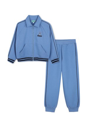 Ensemble de survêtement bleu clair avec passepoil noir, veste zippée avec poches devant, col et petit personnage brodé au-dessus du nom de la marque sur la poitrine.