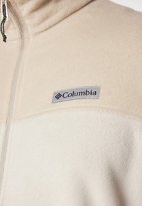 Beige fleecejakke med grå Columbia-logo; har en høj krave og lynlås; blød tekstur og varm optræden.