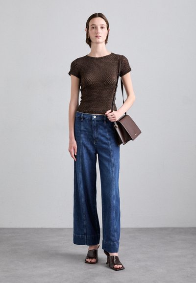 Hnědý pletený top s krátkými rukávy, kombinovaný s širokými modrými džíny s vysokým pasem a výrazným ohybem. Hnědá kožená kabelka a podpatky.
