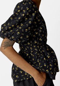 Blusa nera floreale con maniche a sbuffo, vita arricciata e intricati motivi di fiori gialli. Il tessuto testurizzato aggiunge profondità.