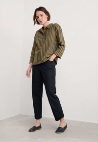 Camisa a rayas en mostaza y azul turquesa, con mangas de tres cuartos, combinada con pantalones de mezclilla oscuros y zapatos negros de slip-on. Fondo liso.