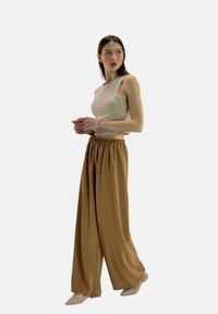 Top senza maniche beige abbinato a pantaloni larghi di colore marrone chiaro con vita elasticizzata. Modella che indossa scarpe di colore neutro, in piedi contro uno sfondo bianco.