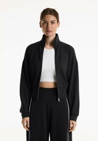 Schwarze, kurz geschnittene Zip-Jacke mit hohem Kragen, langen Ärmeln und glatter Textur, kombiniert mit passenden schwarzen Hosen, die weiße Seitenstreifen haben.