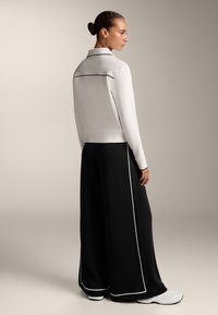 Fehér, galléros, hosszúujjú felső, fekete díszítéssel. Laza fekete nadrággal párosítva, fehér részletekkel. Sima textúrájú anyag.