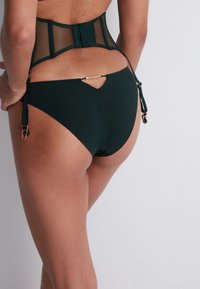 Bas de bikini vert foncé avec une découpe en V, des lanières latérales ajustables avec des éléments en or, et un tissu lisse avec un léger éclat.