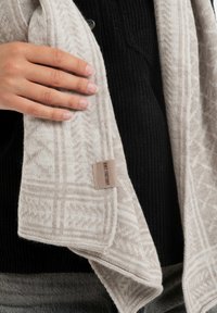 Lichtbeige gebreide sjaal met een geometrisch patroon, zachte textuur en een label aan de rand. Draag over een zwart geribbeld kledingstuk.