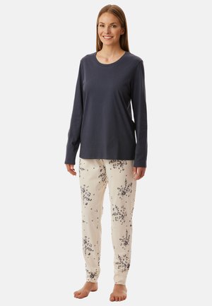 Donna in piedi a piedi nudi, indossa un top scuro a maniche lunghe e pantaloni del pigiama chiari con motivi floreali scuri.