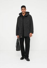 Black, hooded jacket with a button closure, side pockets, and a slightly textured fabric, worn over a black shirt and trousers.