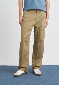 Pantaloni cargo din bumbac bej cu o croială lejeră, având buzunare mari pe față și detalii de cusătură vizibile, asortați cu adidași albi.