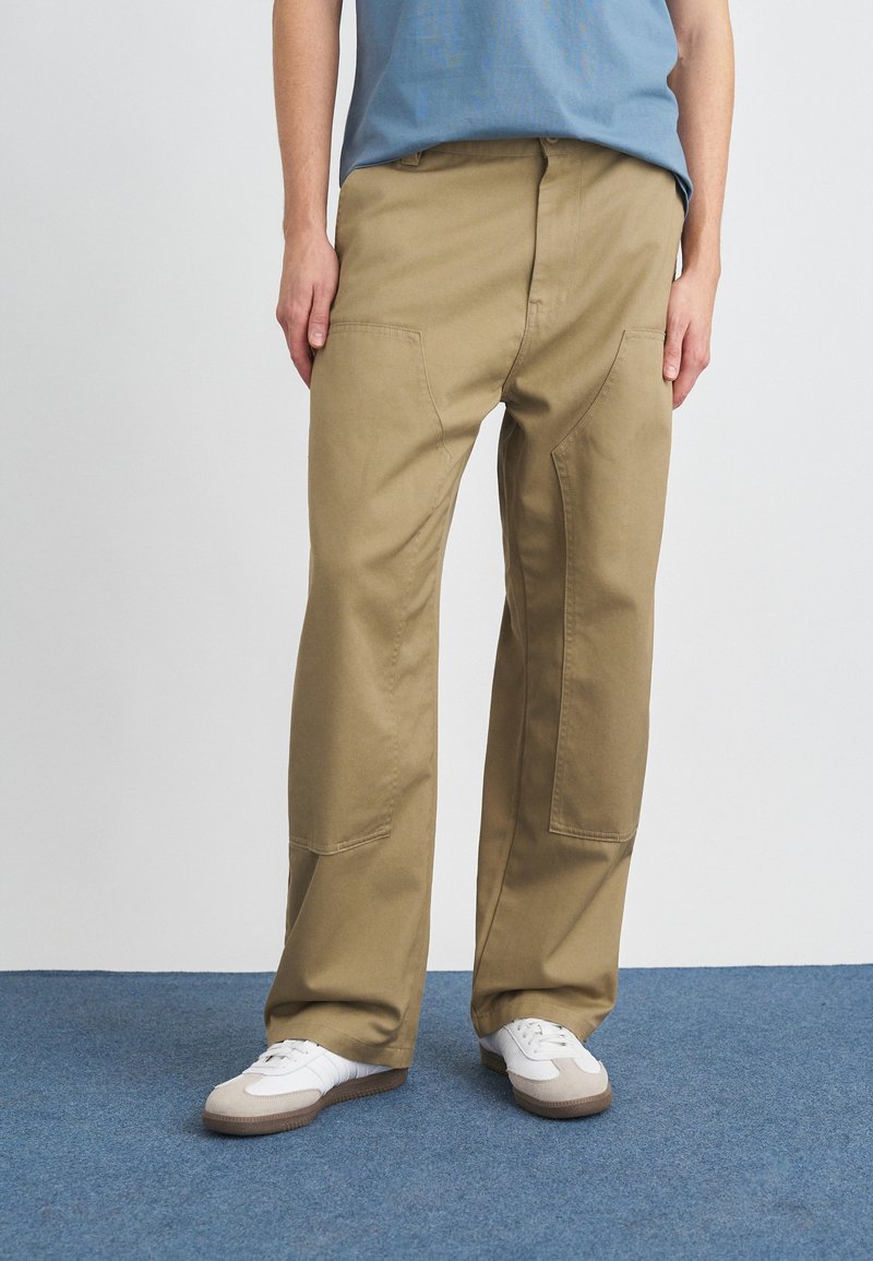 Beige bomullscargobyxor med en avslappnad passform, utrustad med stora framfickor och synliga sömdetaljer, som matchas med vita sneakers.