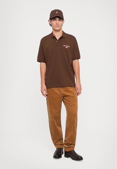 Jovem vestido com um boné e camisa Polo Sport castanhos, calças de veludo cotelê castanho claro com remendos e botas castanhas escuras, de pé contra um fundo branco.