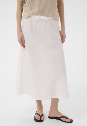 Žena má na sebe bielu midi sukňu so sťahovacou šnúrou v páse, béžový pletený top a čierne sandále na plochej podrážke, stojí pred jednofarebným pozadím.