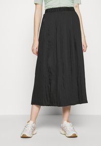 Černá plisovaná midi sukně s elastickým pasem, hladkým povrchem a volným designem, doplněná o světlé tenisky na neutrálním pozadí.