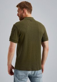 Maslinasto zelena polo majica s mekanom teksturom, kratkim rukavima i zakrpom na lijevom rukavu. Ima klasičan ovratnik i mali logo na stražnjoj strani.