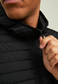 Chaqueta negra acolchada con cuello alto, cierre frontal con cremallera y cordón ajustable. Primer plano muestra una mano agarrando la cremallera.