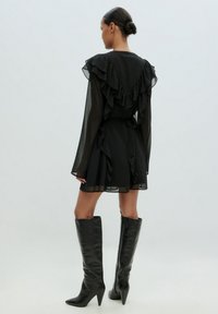 Schwarzes, durchsichtiges Kleid mit Rüschendetails, langen Ärmeln und taillierter Passform, kombiniert mit knielangen schwarzen, spitzen Stiefeln. Sichtbare Pünktchenstruktur.