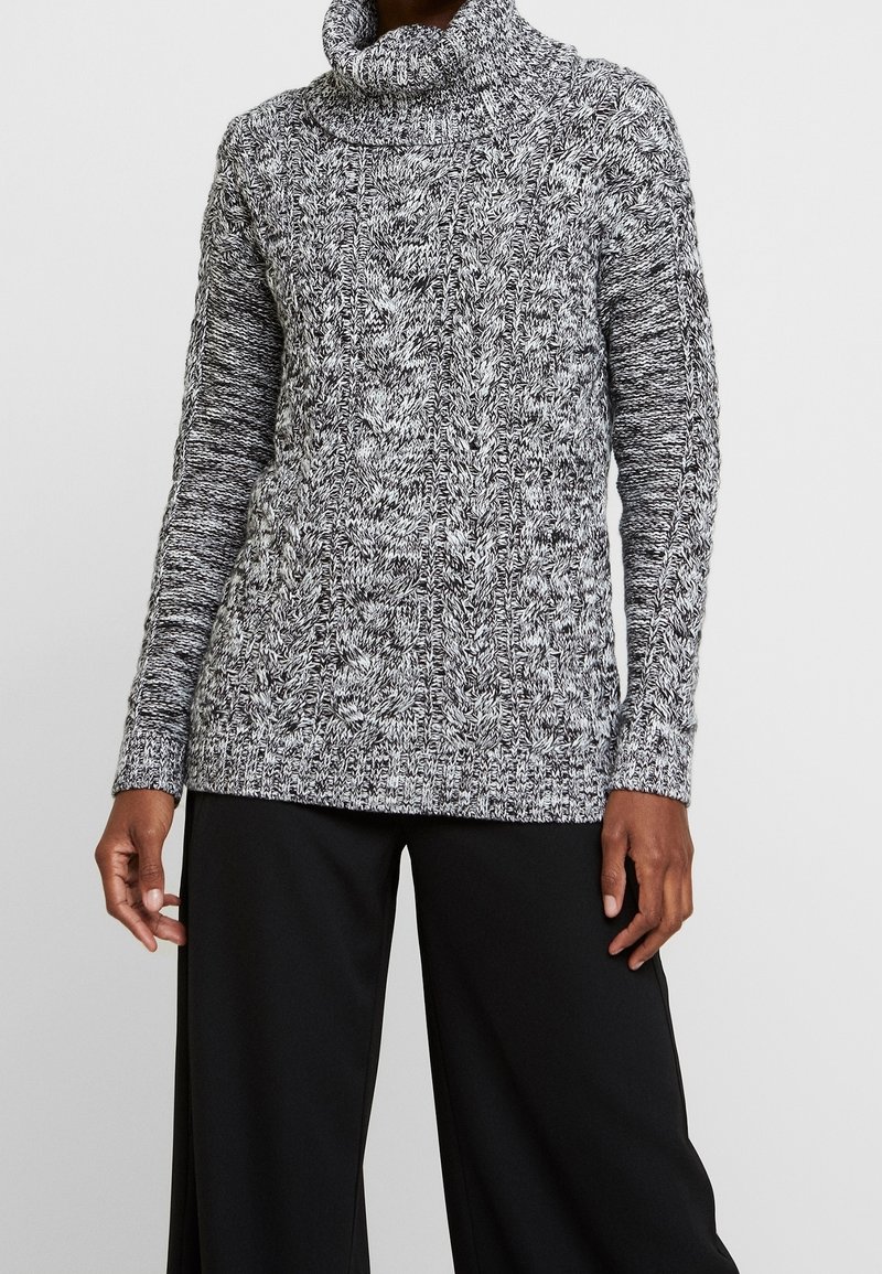 Frau in einem schwarz-weißen, grob gestrickten Rollkragenpullover und schwarzen, weiten Hosen, die vor einem schlichten Hintergrund steht.