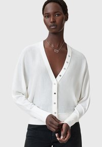 Valkoinen neuletakki, jossa on syvä V-pääntie, napitettava etuosa, ribattu hihansuu ja sileä rakenne. Varustettu hopeanvärisillä napilla etuosassa.