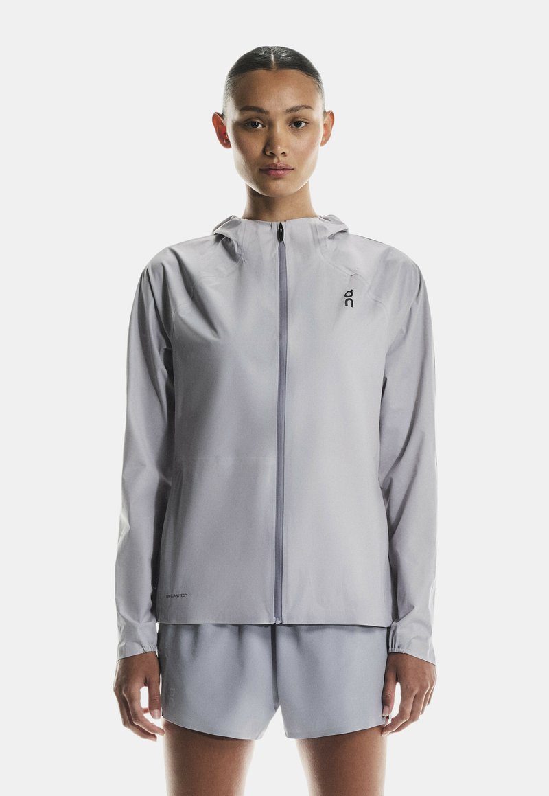 Svetlo sivi jakna s polnim zadrgo, nastavljivo kapuco in stranskimi žepi. Izdelana iz gladkega, vodoodpornega blaga, ima logo na levi strani prsnega dela.