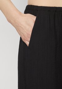 Černá texturovaná látka s detailem kapsy. Pás je elastický a látka má jemný zmačkaný efekt, což zvyšuje její vizuální zajímavost.