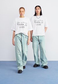 Two models wear loose-fitting white t-shirts with black text and light blue baggy jeans. They stand on a blue floor in a minimalistic setting.