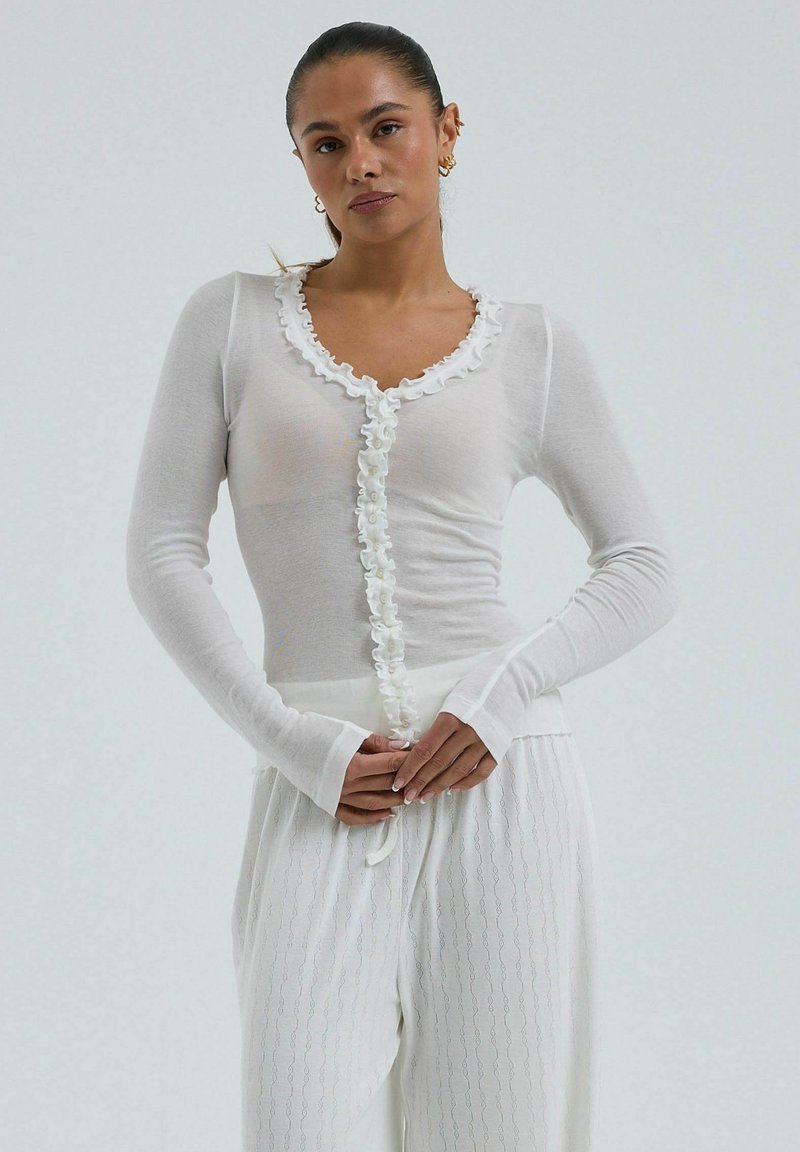 Femme portant un haut blanc ajusté à manches longues avec volant à l'avant et un pantalon blanc à motifs, debout devant un fond clair uni.