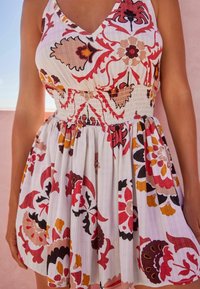 Robe à imprimé floral blanche, avec des motifs rouges, bruns et jaunes. Taille smockée, décolleté en V, et tissu léger avec texture.