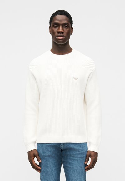 Pull en tricot blanc à encolure ronde, texture côtelée, et logo subtil sur la poitrine, associé à un jean en denim bleu.