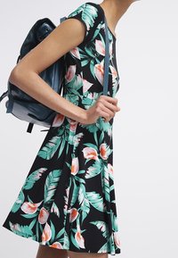 Kobieta w czarnej sukience w zielone i różowe kwiaty, nosząca ciemnoniebieski plecak na jednym ramieniu.