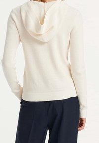 Felpa in misto cotone di color bianco crema, con texture a coste, un ampio cappuccio e polsini aderenti, abbinata a pantaloni scuri.
