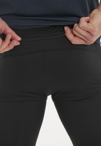 Mustat urheilulegginsit, joissa on sileä pinta ja huomaamaton vetoketjullinen tasku takana, jota henkilö säätää vyötärön kohdalta.