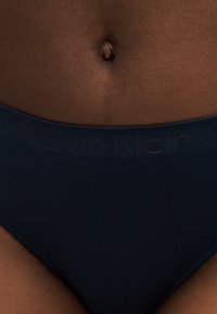 Κοντινό πλάνο σκουρόχρωμης κοιλιάς που φοράει εσώρουχα Calvin Klein σε ναυτικό μπλε χρώμα με εμφανές λογότυπο στη μέση της ζώνης.