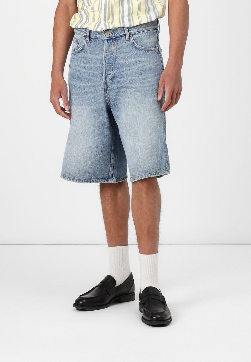 Light wash denim shorts with a loose fit, raw hems, and classic five-pocket styling. Paired with black loafers and white socks.