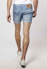 Shorts de mezclilla en azul claro con un diseño desgastado, puños enrollados y un parche de cuero marrón de la marca en la parte trasera de la cintura. Calzado casual visible.
