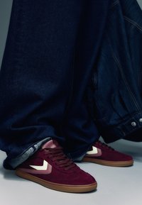 Baskets en daim bordeaux avec détails blancs, dotées d'une semelle en gomme. Portées avec un jean denim foncé, légèrement retroussé aux chevilles, révélant les détails des chaussures.