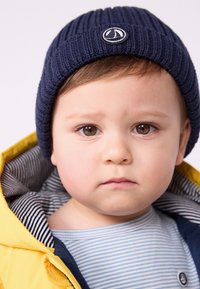 Berretto a coste navy con logo, indossato da un bambino. Giacca gialla con foderatura a righe. Caratteristiche facciali degne di nota includono occhi marroni.