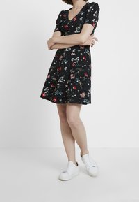 Černé šaty s krátkým rukávem, zdobené květinovým potiskem s červenými a růžovými květy, s výstřihem do V a rozšířenou sukní, doplněné o bílé tenisky.