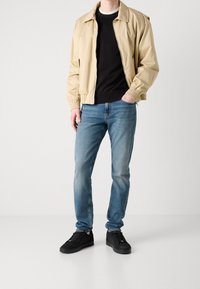 Beige bomber jacket with a black sweater underneath, paired with light blue jeans and black sneakers. Soft fabric, elastic cuffs on jacket.