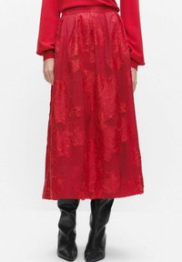 Red floral-patterned maxi skirt with a pleated design and a sheer overlay. Features a high waist and textured fabric. Paired with black boots.