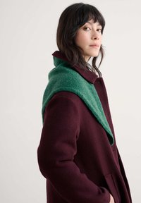 Burgundy frakke med en struktureret overflade, der har en grøn, strikket omslagskrave. Designet inkluderer en enkelt knaplukning og sidelommer.