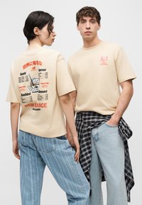 Deux mannequins portent des t-shirts beige New Balance ; l'un montre l'avant avec un petit graphique et l'autre montre le dos avec un imprimé de menu sur le thème du sushi.