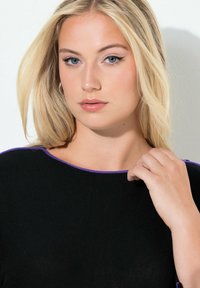 Czarny prążkowany top z okrągłym dekoltem ozdobionym fioletowymi akcentami. Modelka ma długie blond włosy i delikatnie dotyka kołnierza.