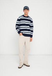 Maglione a righe navy e bianche a maglia con scollo rotondo a coste, abbinato a pantaloni in corduroy beige chiaro a gamba larga e sneakers beige.