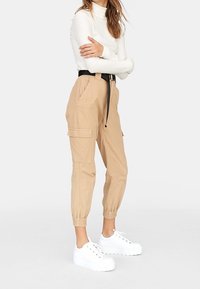 Beige cargobroek met elastische boorden en riem, voorzien van zijzakken. Gedragen met een aansluitend wit shirt en stevige witte sneakers.