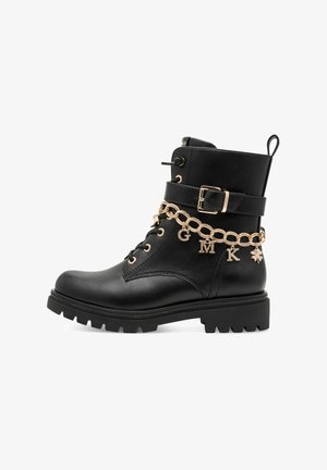 Botte de combat en cuir noir avec sangle à boucle dorée et chaîne décorative dorée avec breloques lettres G, M, K et un trèfle.