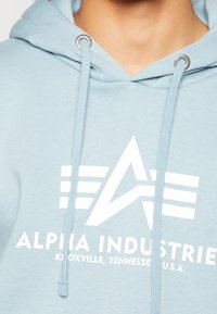 Felpa con cappuccio azzurro chiaro in tessuto morbido, con logo grafico bianco e testo, dotata di coulisse abbinata e occhielli in metallo.