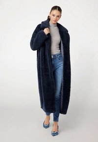 Manteau long en fausse fourrure bleu marine avec un grand col, porté sur un haut gris et un jean bleu. Dispose d'une doublure à pois et d'une texture douce.